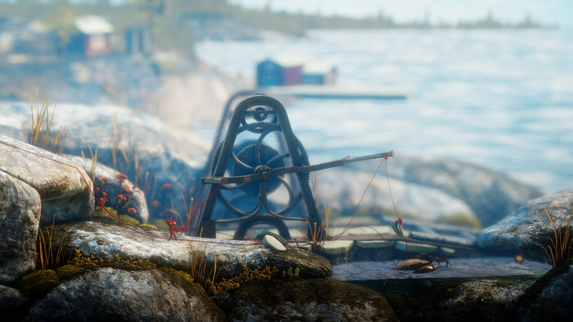 A video game picture of a little red yarn figure, pulling a red string through an iron reel, on rocks by water. 