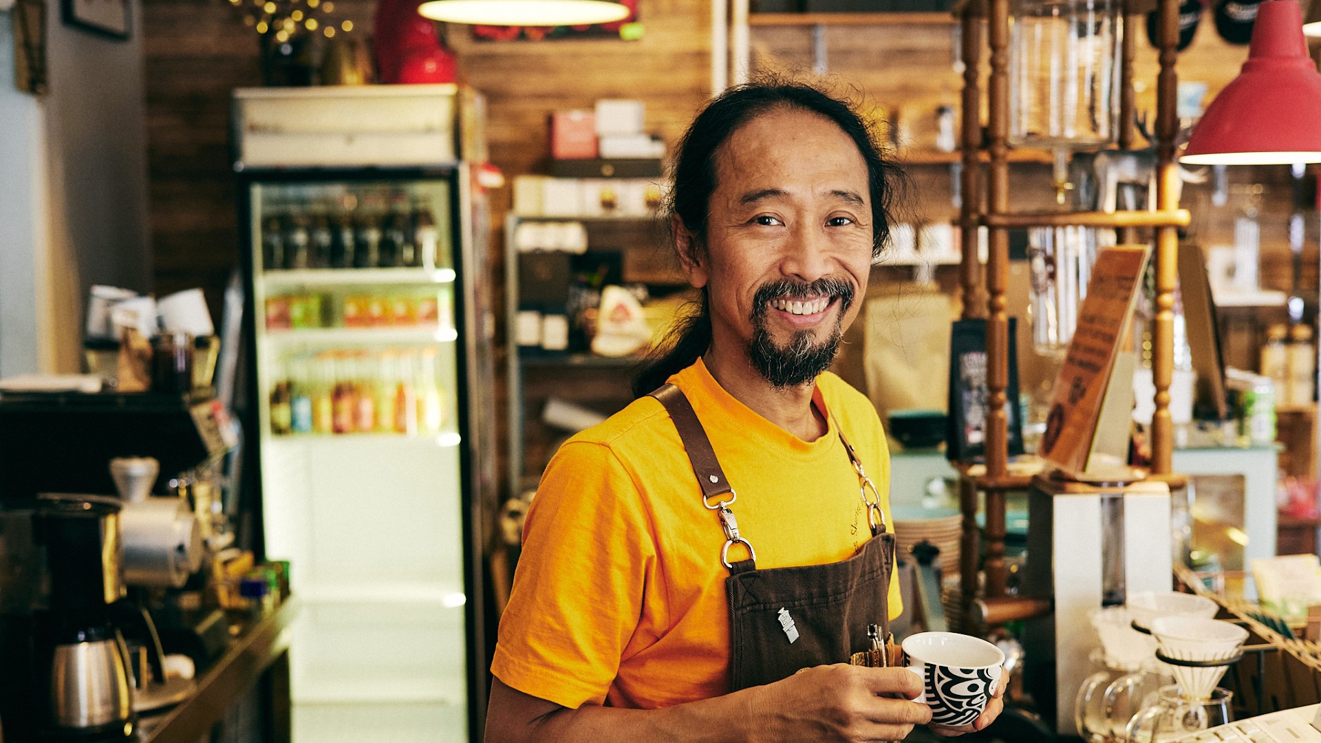 A man in a yellow sweater holding a coffee cup, smiling towards the camera. 