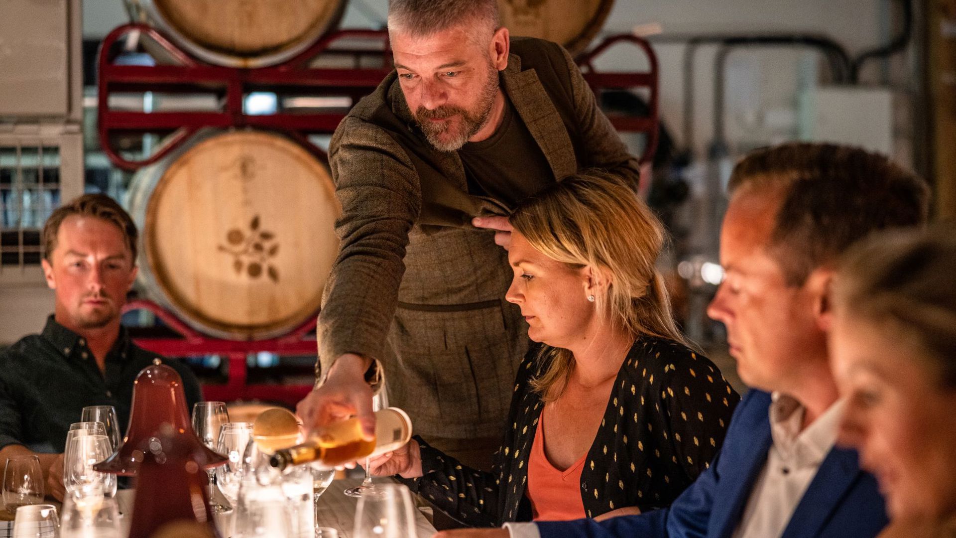 A man pouring cider in a womans glas, suppounded by casks. 